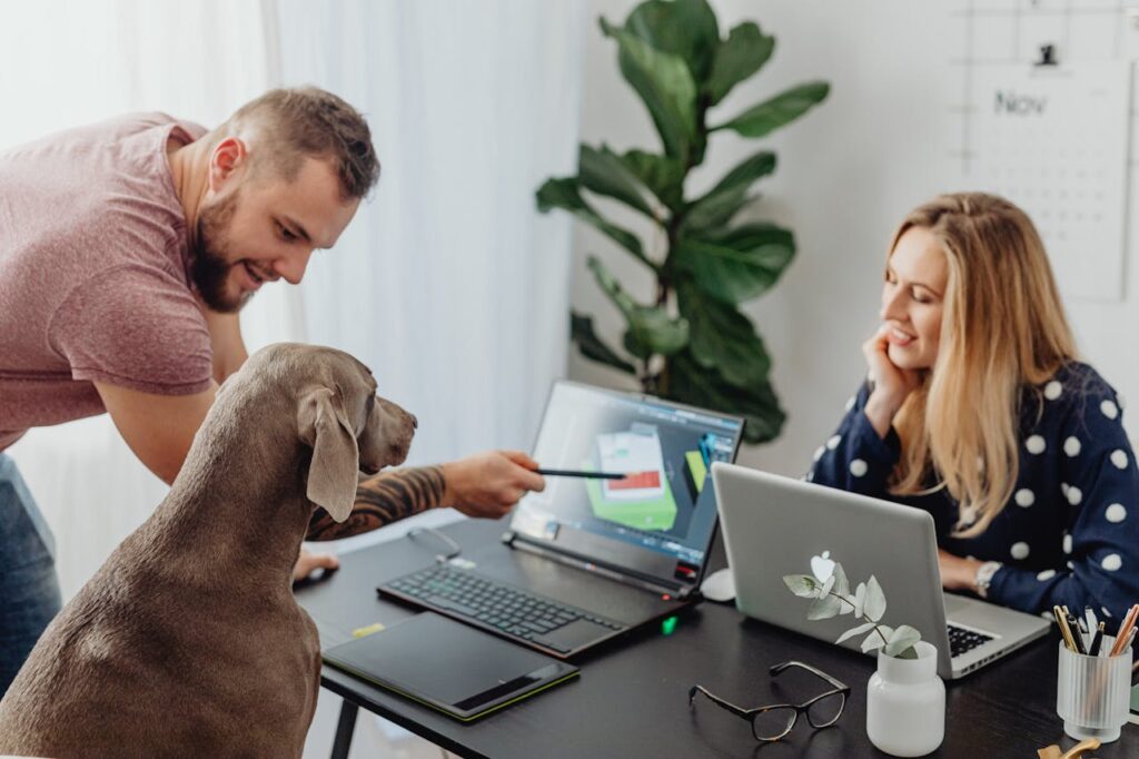 Cane seduto alla scrivania davanti a un computer portatile mentre due colleghi osservano divertiti